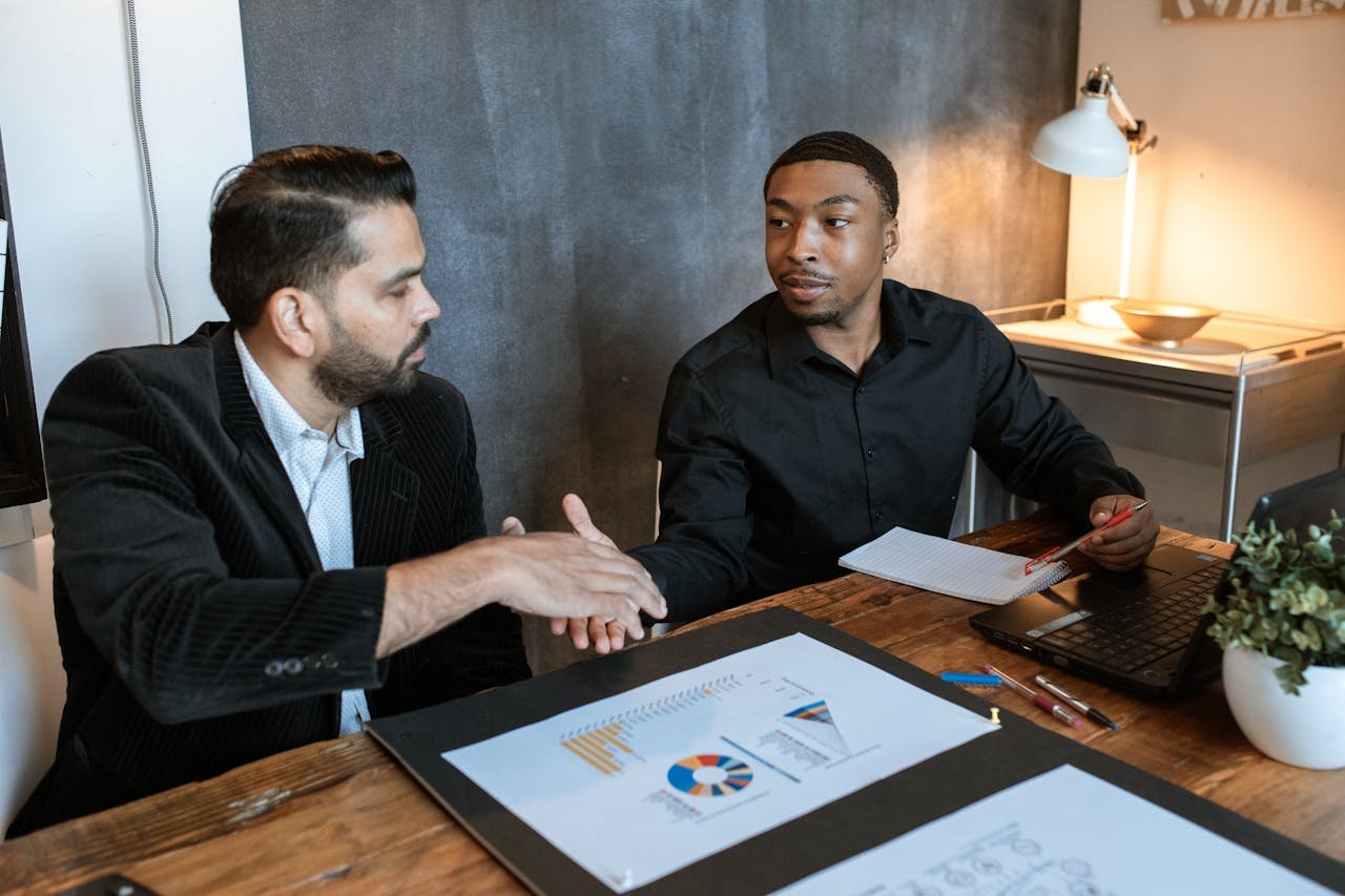Two businessmen shaking hands over a desk with charts in a modern office.